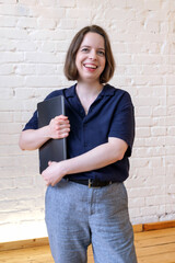 Vertical portrait of young brunette woman with shoulder length hair on white brick wall background. Woman in blue shirt stands with laptop in her hands and smiles. Concept of passionate student life