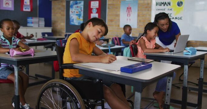 Mixed race schoolboy in wheelchair, sitting in classroom making notes, teacher in background