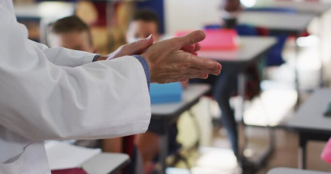 Diverse teacher disinfecting hands in classroom, with schoolchildren sitting wearing face masks