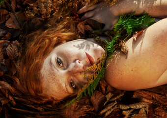Beautiful sexy lovely young redhead girl lying on red golden autumn leaves, with a frond of fern on his chin, in the forest, with friendly seductive smile in the face