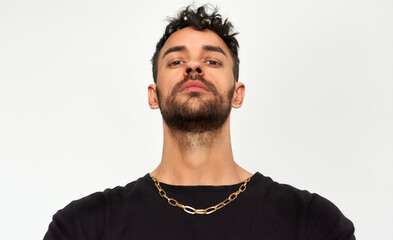 Portrait of a confident serious young man wearing black t-shirt and a gold necklace, looks directly at camera, isolated over grey studio background.