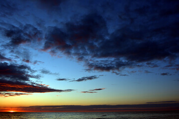 The twilight blue-scarlet sky at sunset with clouds, which turned from light to black, over the calm sea, the edge of the setting sun can be seen on the horizon.