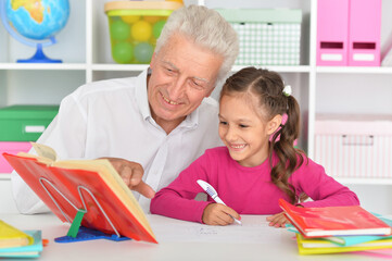 Cute girl doing homework with grandfather at the table at home