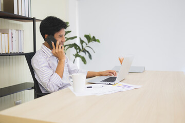 Young Asian businessman is calling with a smartphone and using a laptop and smiling in the office. start-up and business concept. stay home. work from home.