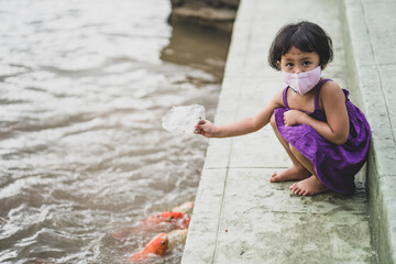 Adorable Asian girl wearing healthy face mask feeding koi carps at pond.