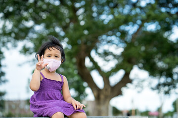 Adorable Little girl wearing protective face mask sitting against tree outdoor.