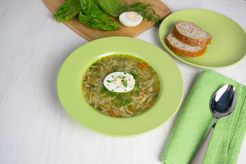 Noodles with chicken broth, egg, spoon and dill in green plate on white background