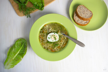 Noodles with chicken broth, egg, spoon and dill in green plate on white background