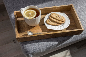 A white cup of tea with lemon, a long cupronickel spoon with a twisted handle and a saucer with three oatmeal cookies for breakfast on a wooden tray, a rag napkin on a gray sofa.