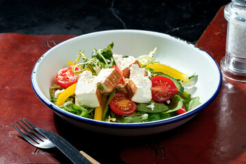 Salad with smoked feta, cherry tomatoes, bell pepper and spinach in a white plate. Dark background. Hard light