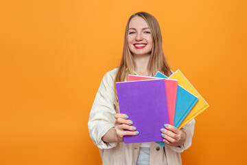 Caucasian blonde student girl holds four books in multi-colored covers smiling isolated over orange background in studio. English language school, education concept