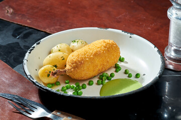 Chicken Kiev with peas, potatoes and green sauce in a white plate on a dark background. Hard light. Deep-fried cutlet