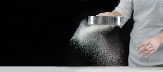 Woman sifts flour through a sieve on a wooden table. Isolated on dark background