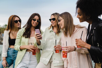Group of girls watching social network content on the smartphone screen drinking fruit cocktails...