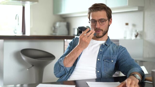 Smiling young man recording voice message on smartphone. Handsome guy using virtual assistant app sitting at home. Voice recognition, audio messages, using translator on mobile phone.