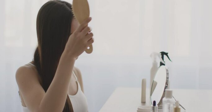Young middle eastern lady combing her her hair, looking at mirror and smiling, sitting at dressing table at bedroom