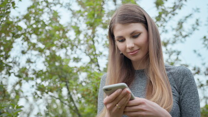 Happy woman typing by mobile phone outdoors. Cheerful girl with smartphone in park on a background. Smiling lady holding cellphone in hands outside. Sport Girl, internet technology, runner exercise