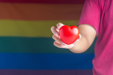 Hand holding a red heart against the background of the rainbow flag (LGBT). Close-up photo. Love concept