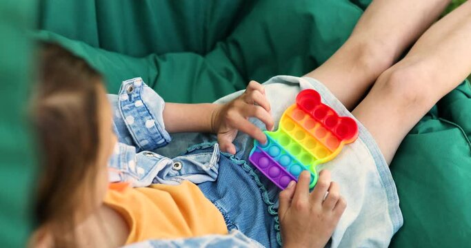 A little girl the on hanging chair outdoors play pop it, kid hands playing bubbles of a rainbow destressing toy, fidget toy in backyard of house on a sunny summer day, summer time vacation.