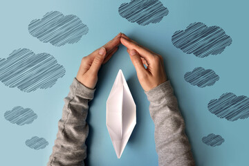 Hands protect the paper boat from bad weather. A symbol of protection of life and an easy way in life - obrazy, fototapety, plakaty