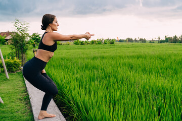 Barefoot female yogi in black tracksuit practice hatha sport near rice fields in Thailand keeping...