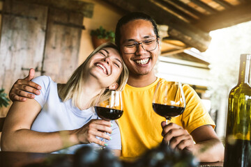 Young couple in love on a date having fun drinking red wine in farmhouse garden - Multiracial...