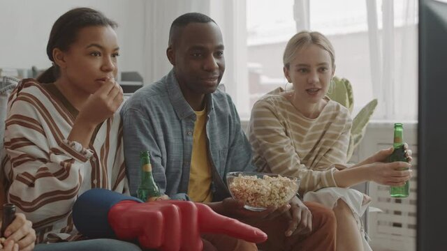Backlighted of African man, Mixed-Race and Caucasian woman sitting on couch in living room, drinking beer and eating snacks, watching favourite sports on television