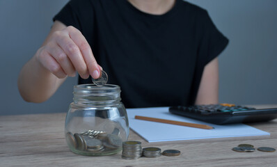 A woman puts a coin in a jar on a wooden table to save money And record household income on the tablet.