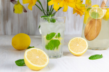 glass of lemon lemonade with mint and ice on white background, yellow flowers in jar