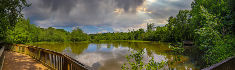 Obraz na płótnie Canvas a stunning panoramic shot of the vast lake water with lush green trees and plants reflecting off the lake with blue sky and clouds on the Doll's Head Trail at Constitution Lakes in Atlanta Georgia