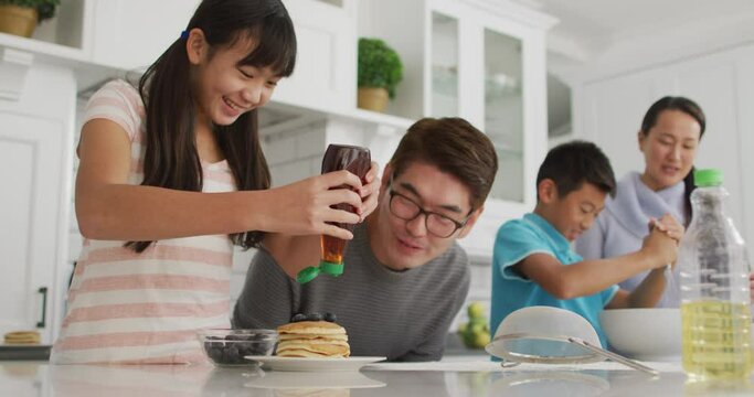 Happy asian parents in kitchen cooking with son and daughter, daughter putting syrup on pancakes