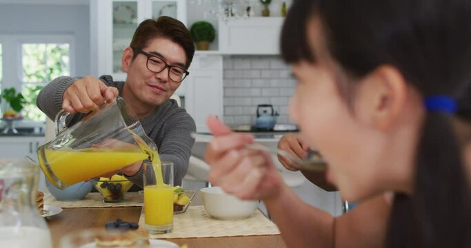 Happy asian father in kitchen having breakfast pouring juice for smiling son and daughter