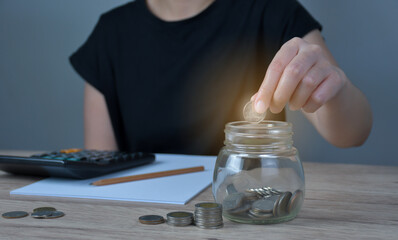 A woman puts a coin in a jar on a wooden table to save money And record household income on the tablet.