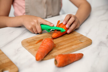Little girl peeling carrot at table in kitchen, closeup. Preparing vegetable
