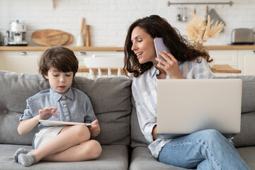 Caring mom businesswoman talk to small son during business phone call as work on laptop computer at...