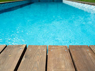 Ripped wavy water surface in swimming pool with wooden, old deck and green grass around. Swimming pool area in hotel or villa. Summer vacation background.