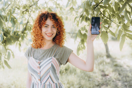 Redhead girl with afro curls with a phone in a green park makes a selfie and smiles, makes a smartphone presentation