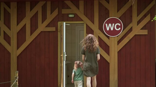 woman and girl go to public toilet. Observance of gi on street in a wooden building. Mother and daughter hold hand walk to wc while walk. Parents and children together in happy family concept. 