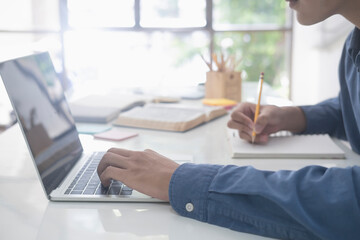Student studying and writing notes in a notebook at home, making research on laptop and browse...
