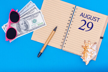 Travel concept flat lay - notepad with the date of 29 august pen, glasses, dollars and seashell on blue background.