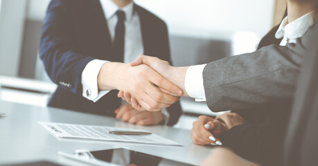 Unknown businessman shaking hands with his colleague or partner above the glass desk in modern office, close-up. Business people group at meeting