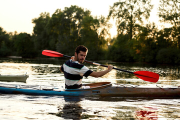 Enthusiastic young caucasian guy holding a paddle while boating on a lake surrounded by nature