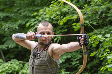 Portrait of a stern man shooting an arrow from a bow in the forest. Concept: men's hobbies,...
