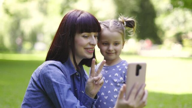 Pretty 30-aged happy mother with her daughter taking selfie picture in the park, smiling and having fun, showing victory peace sign to smartphone camera. People, technologies concept
