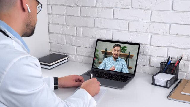 Doctor wears lab coat in hospital office writes symptoms in notebook listen patient online video conference call webcam chat laptop. Smiling man client in computer screen tells remote medical worker