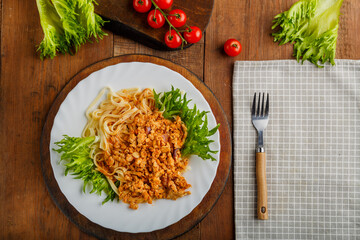 A plate of bolognese pasta decorated with greens on a wooden table next to a gray napkin and a fork and cherry tomatoes.