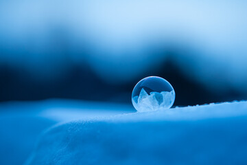 A frozen soap bubble with growing ice crystals and shallow selective focus on a blue winter background and deep snow