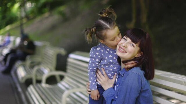 Close-up, video stock footage. Slow motion soft focus. Pretty young Caucasian mom hugs her cute little daughter, sitting on the bench in the park. Adorable girl kisses her mommy on the cheek