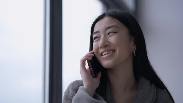 Close-up portrait of positive Asian young woman with brown eyes talking on the phone in the morning at window laughing. Cheerful female freelancer discussing work plan in home office indoors
