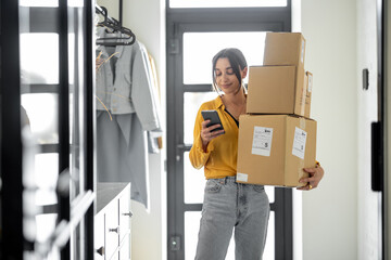 Young woman with parcels at the hallway at home, holds smart phone and checking goods. Concept of buying online and delivering goods home - 438671614